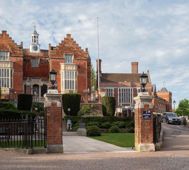 A large historic brick building with multiple gabled roofs and tall, ornate windows, set behind a gated entrance with brick pillars topped by lantern-style lights. The entrance features a paved pathway leading up to the building, flanked by well-maintained green lawns, trimmed topiary bushes, and decorative stone elements. The setting appears to be in daytime with natural light illuminating the scene and a cloudy sky overhead. This property exemplifies a stately home interior or exterior relevant to house removals and relocation services provided by manwithvanharrowonthehill.co.uk, with the exterior view showing mature landscaping and architectural details suitable for furniture transport or home relocation projects.