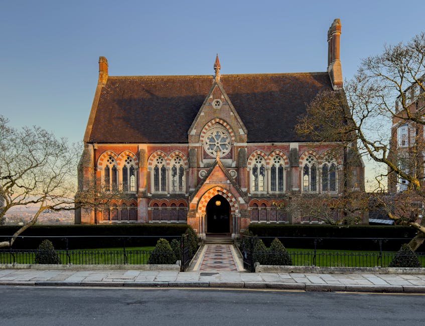 A historic red-brick building with stepped gables and arched windows, situated behind a black wrought-iron fence and a stone balustrade, is visible against a partly cloudy sky. The building features o