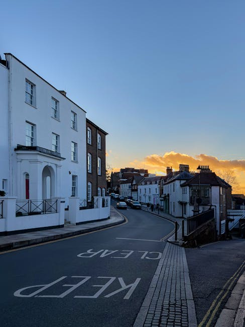 A quiet residential street in Harrow on the Hill during sunset, with a steep incline and a 'KEEP CLEAR' road marking painted on the asphalt. On the left side of the image, there is a large white, multi-story building with tall windows, a small balcony with black railings, and an arched doorway, possibly part of a home or apartment complex. The street curves gently to the right, lined with additional white and dark-colored terraced houses built in a traditional style, featuring tiled roofs and chimneys. A few cars are seen parked along the curb, and the sky above displays orange and golden hues with scattered clouds, indicating the end of the day. The scene captures an environment suitable for professional house removals and furniture transport, with an emphasis on clear access and the residential nature of the area, consistent with the focus on home relocation and moving logistics within Harrow on the Hill, as provided by [COMPANY_NAME].