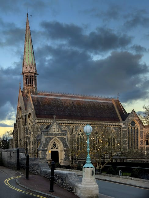 A historic stone church with a tall, pointed spire and intricate Gothic architectural details sits alongside a narrow street in Harrow on the Hill. The church's façade features arched windows with tracery, stone carvings, and a steep, tiled roof. In the foreground, a vintage-style lamp post stands on a small plinth near the curved road, which is lined with black bollards and yellow double lines along the curb. The scene is captured during late afternoon or early evening, with dark clouds overhead and a soft, diffused light illuminating the church. This setting provides a neutral, factual view of the church's exterior, suitable for illustrating content related to house or furniture removals, house relocation, or moving services in the Harrow on the Hill area, as seen on the page about Church Street moves by [COMPANY_NAME].