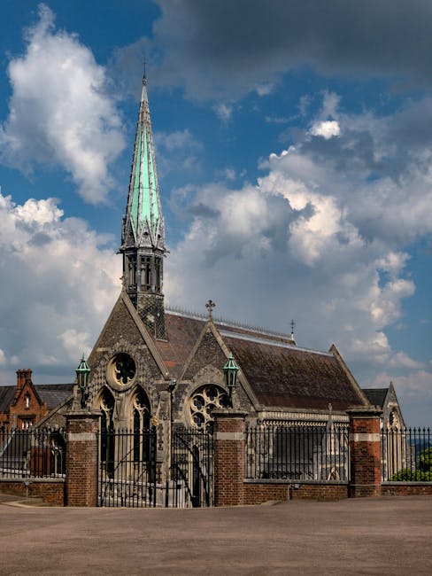 A historic stone church with a tall, pointed spire topped with a weathered copper roof, set against a partly cloudy sky with blue patches. The church features arched windows with intricate tracery, small decorative crosses on the roof ridges, and a wrought iron fence with brick pillars surrounding the perimeter. The surrounding area is paved, with no visible vehicles or people in the immediate vicinity. The church’s exterior presents traditional gothic architectural elements, and the scene appears to be captured during daylight hours. This image could relate to house removals or home relocation services by [COMPANY_NAME], emphasizing careful transportation and logistical planning involved in moving furniture and household items in and around historic properties like this church, which may be part of a relocation or moving process in a residential or community setting.