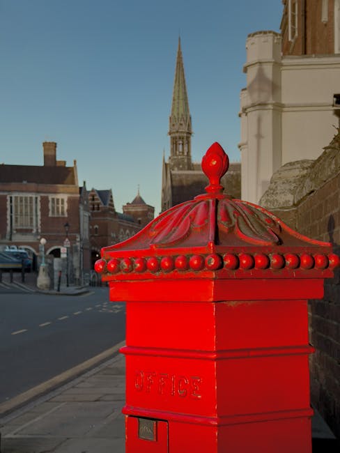 A bright red traditional British post box with a domed, decorative top and rounded bead details along the edge, situated on a sidewalk near an ornate stone building. Behind the post box, there is a street with parked cars and a few pedestrians walking along the pavement. In the background, a church steeple with a pointed spire reaches into a clear blue sky, suggesting a historic town centre setting. The surrounding architecture includes historic brick buildings, and the scene is illuminated by natural daylight. The image captures the urban environment during daytime, reflecting elements typical of a UK town or city centre, suitable for context about home relocations or house removals in Harrow on the Hill, with [COMPANY_NAME] providing moving services.