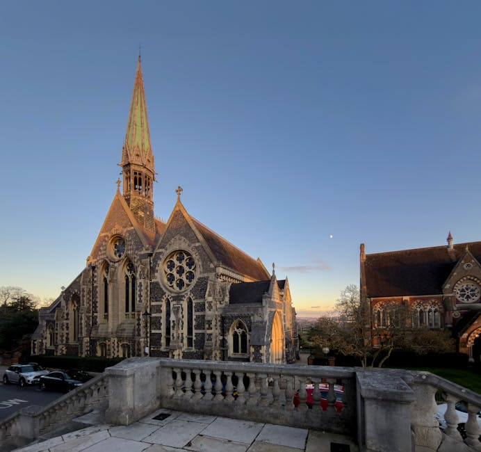 A street scene in Harrow with a historic church featuring a tall, pointed spire and intricate stone architecture, situated on a slightly inclined road. To the left, there is a red car parked along the