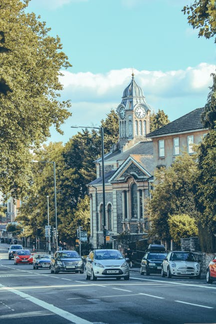 A view of a tree-lined street in Harrow on the Hill during daytime, featuring parked cars along the pavement and a historic clock tower with a domed roof and Gothic architectural details in the background. The street is busy with several vehicles, including sedans and hatchbacks, waiting at a traffic signal. The scene is illuminated by daylight, with a clear blue sky and some scattered clouds overhead. Surrounding buildings include residential houses and small shops, with lush green trees providing shade. This setting illustrates a typical urban environment where home relocation and furniture transport services by [COMPANY_NAME] could be required, especially during moves involving street access and parking considerations.