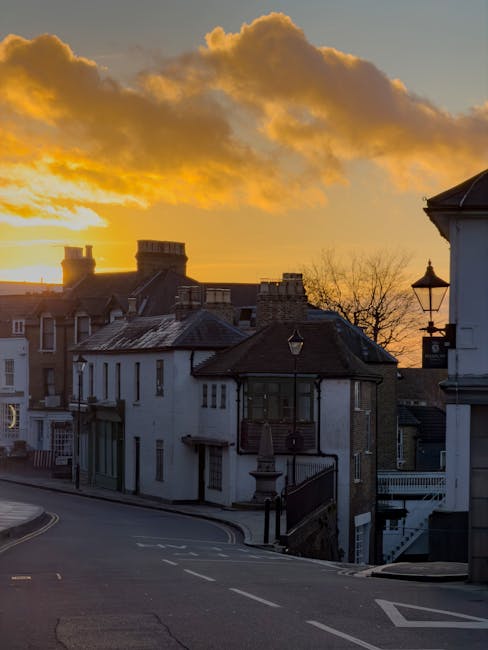 A street scene at sunset in Harrow on the Hill featuring traditional residential buildings with various roof styles, including tiled and flat roofs, some with chimneys. In the foreground, there is a p