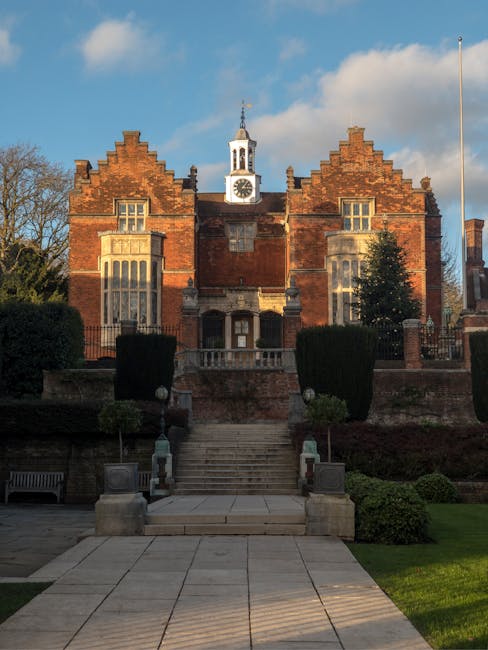 Photograph of a historic red-brick building with a central clock tower featuring a white spire, set against a partly cloudy sky during daytime. In front of the building, there is a paved pathway leading up a short flight of stone steps flanked by low stone walls and decorative stone urns. The building's facade has large, ornate windows with stone detailing, and the surrounding area includes neatly trimmed bushes, small trees, and a park bench on the left side. The scene likely depicts a prominent local landmark or public building in Harrow, which could be relevant for home relocation or moving services by companies like manwithvanharrowonthehill.co.uk involved in house removals, furniture transport, and packing and moving processes.