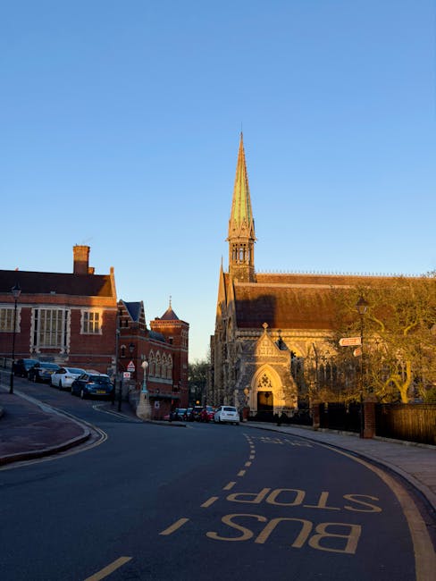 Photograph of a historic red-brick building with a central clock tower featuring a white spire, set against a partly cloudy sky during daytime. In front of the building, there is a paved pathway leading up a short flight of stone steps flanked by low stone walls and decorative stone urns. The building's facade has large, ornate windows with stone detailing, and the surrounding area includes neatly trimmed bushes, small trees, and a park bench on the left side. The scene likely depicts a prominent local landmark or public building in Harrow, which could be relevant for home relocation or moving services by companies like manwithvanharrowonthehill.co.uk involved in house removals, furniture transport, and packing and moving processes.